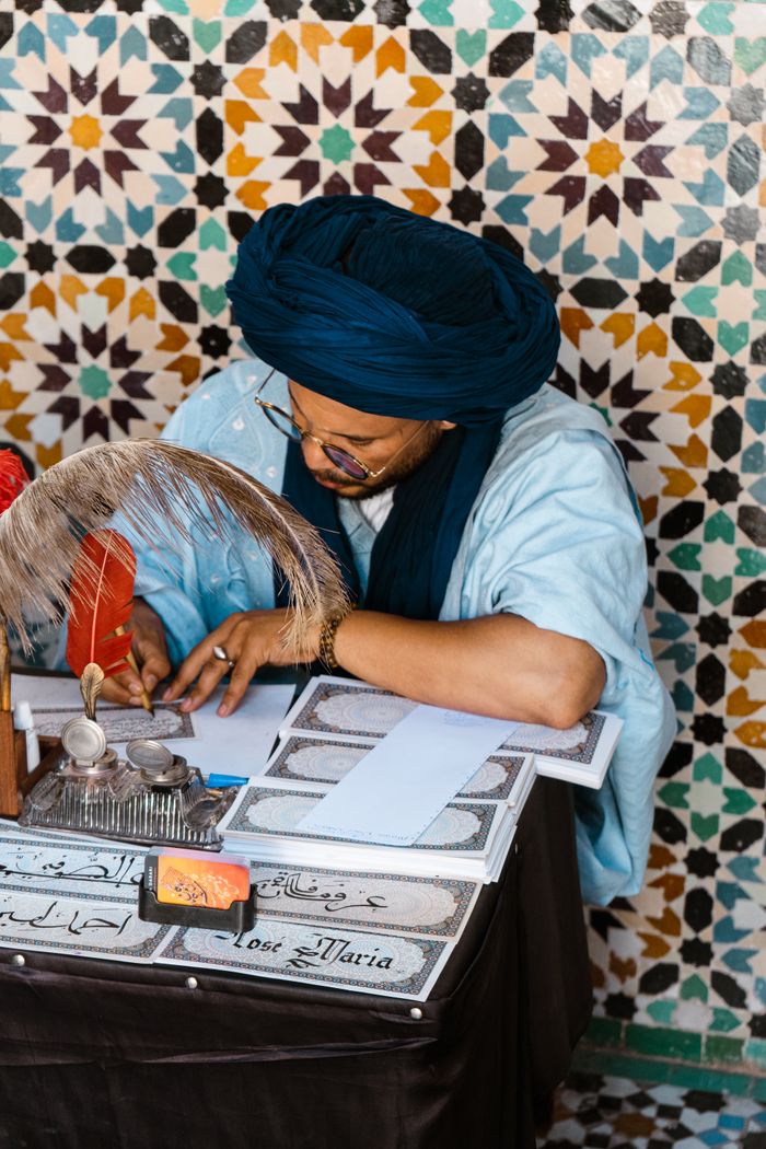portfolio-06 A Moroccan calligrapher writes using traditional tools against a mosaic wall.